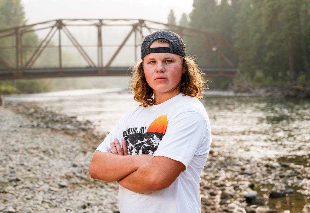 Cle Elum Senior Photographer male student posing with arms crossed, a bridge and river behind him