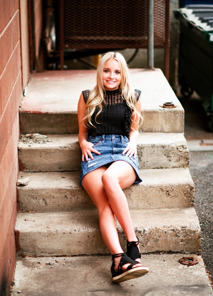 Senior Photography in Ellensburg a young blonde high school student smiles at the camera wearing a skirt and black top sitting on concrete steps