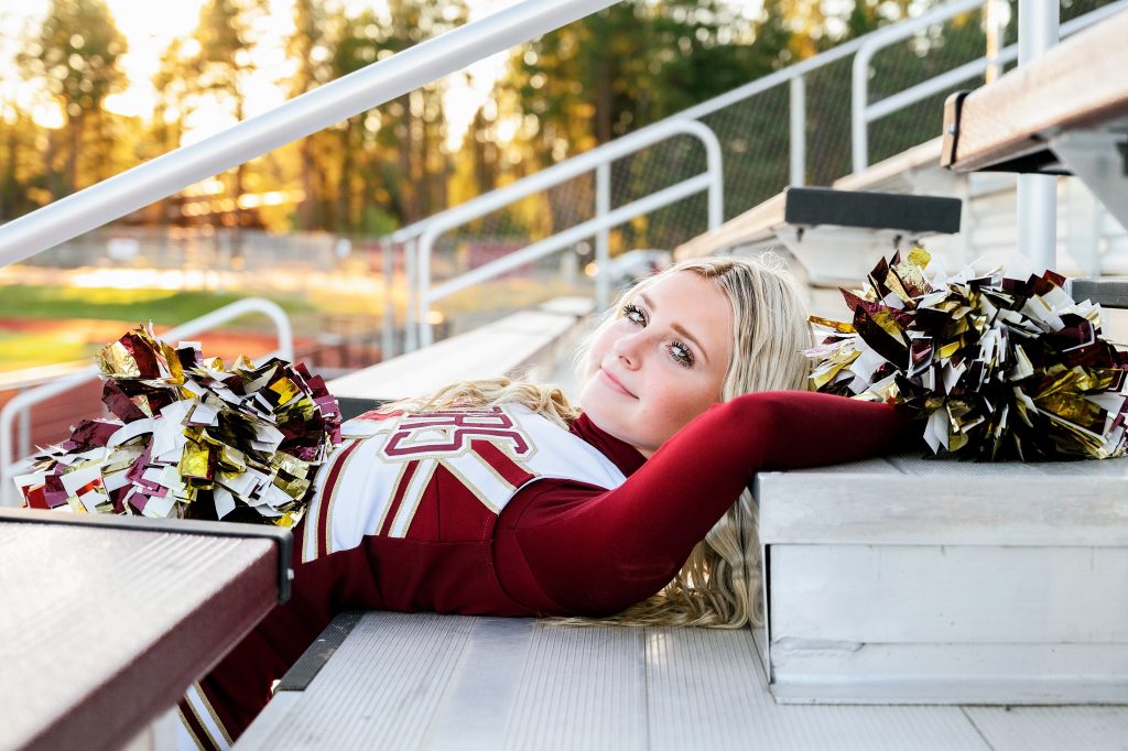 Senior Photography in Ellensburg, a high school girl with blonde hair wearing a cheer leader uniform posing on bleachers with her pom poms 