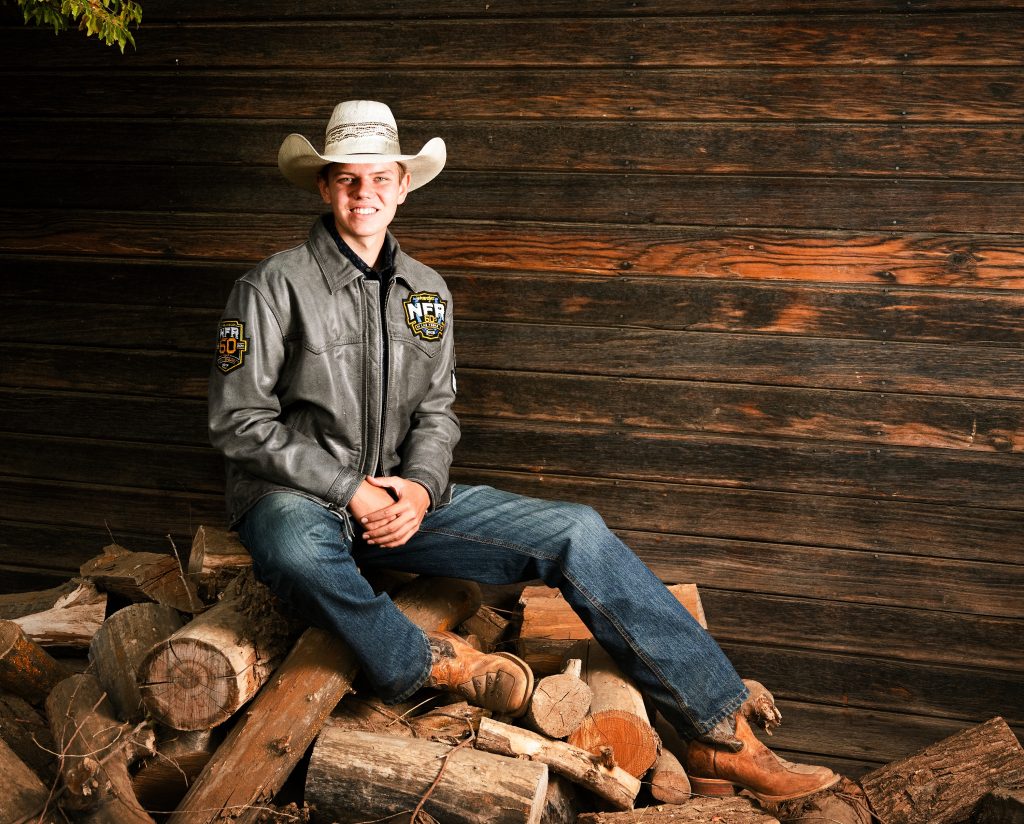 Senior Photos in Ellensburg student sitting on a pile of chopped wood with his rodeo jacket and cowboy hat