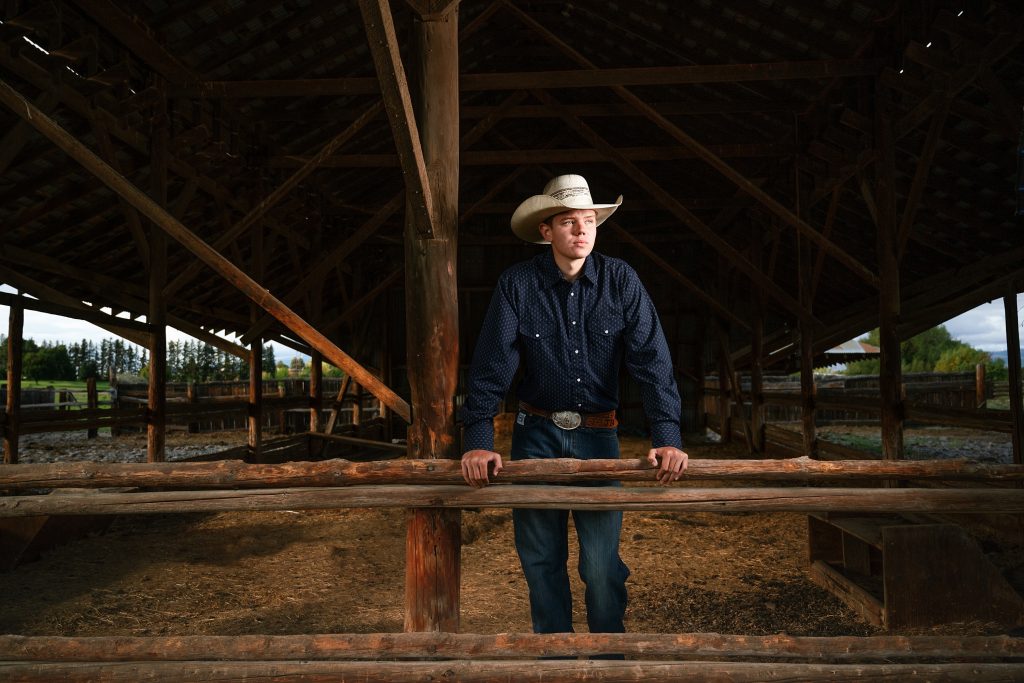 Senior Photos in Ellensburg fourth generation rodeo ranger and student leans against railing wearing classic jeans, belt and dress shirt with cowboy hat