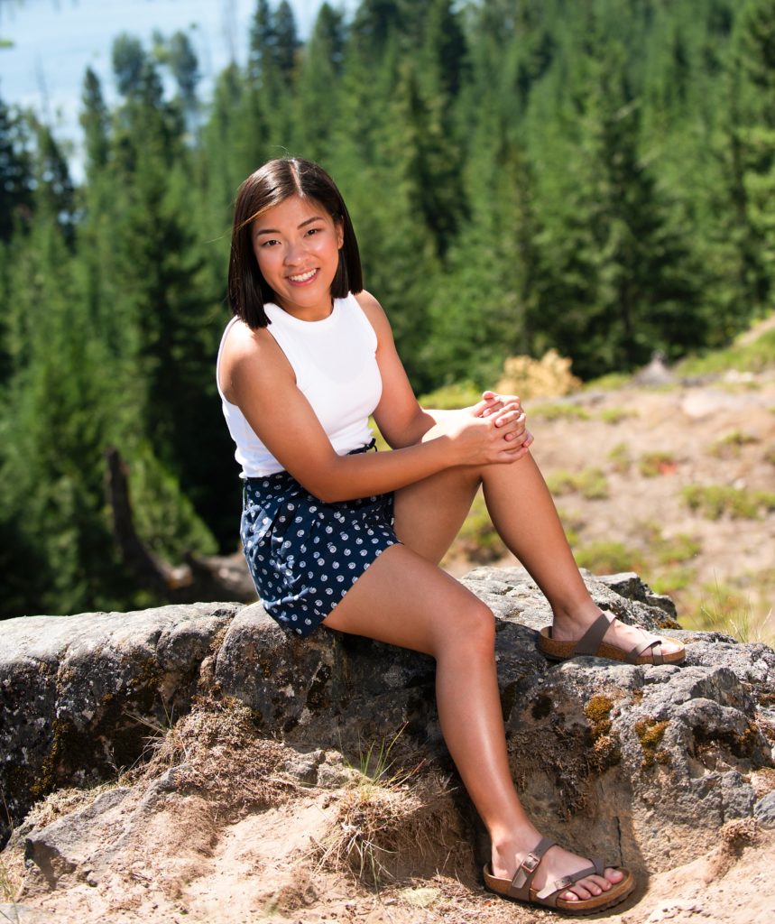 High school senior girl sit on a rock on a mountain side in shorts and tank top and smiles at the camera Suncadia Senior Photographer