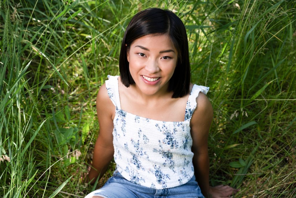 High school senior girl sit in grass and smiles at the camera wearing a white floral top with ruffles in Suncadia