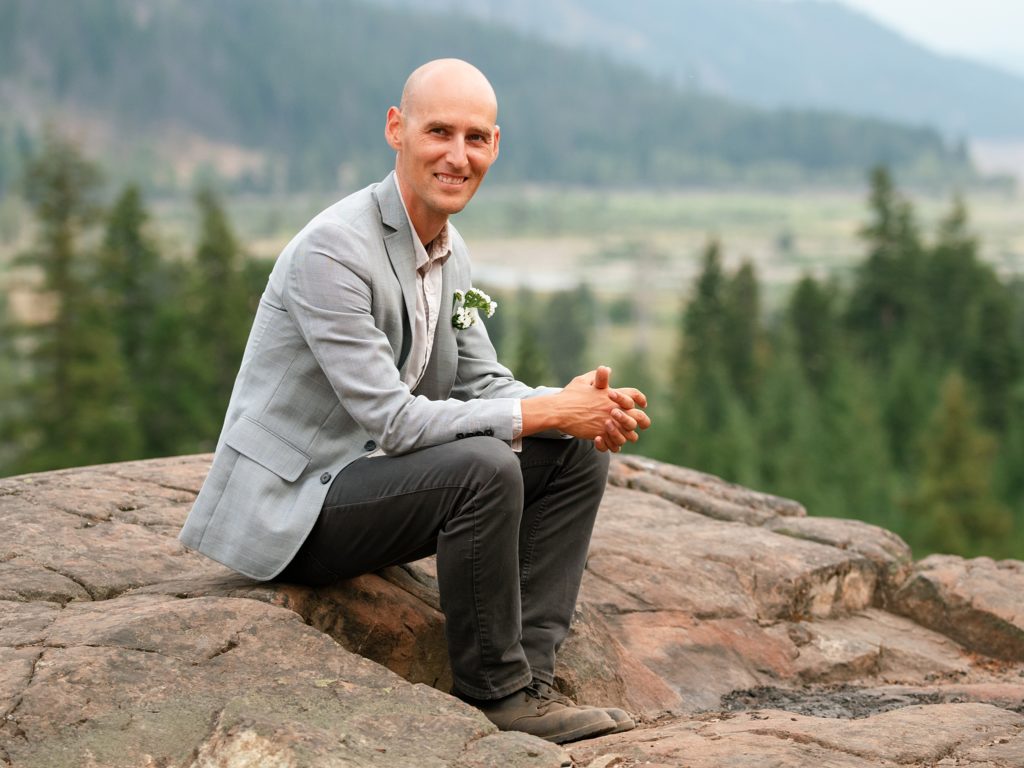 Groom sits on a rocky outlook smiling at the camera with forest in the background in Cle Elum