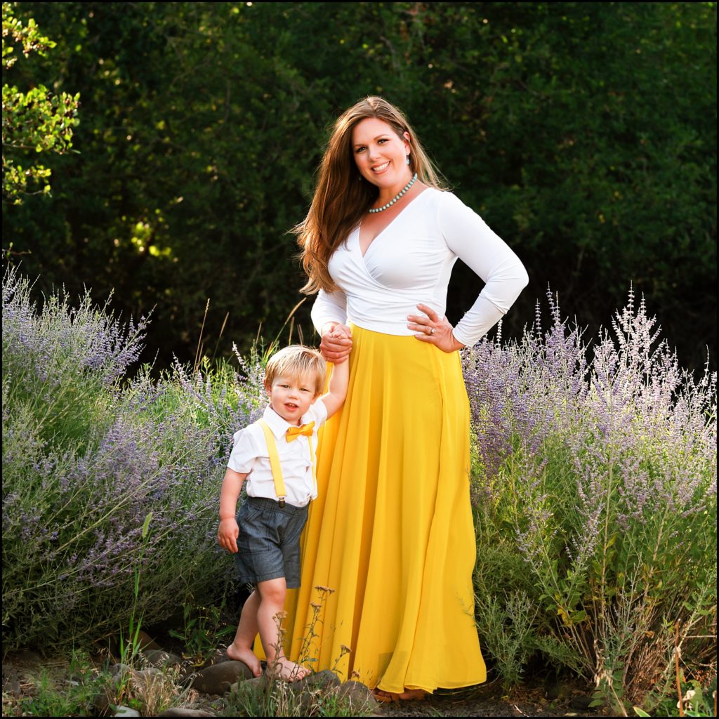 Little blonde boy in white shirt and bow tie holds his moms hand in a field of wild flowers. She is wearing a yellow wrap dress. Ellensburg Family Photographer.