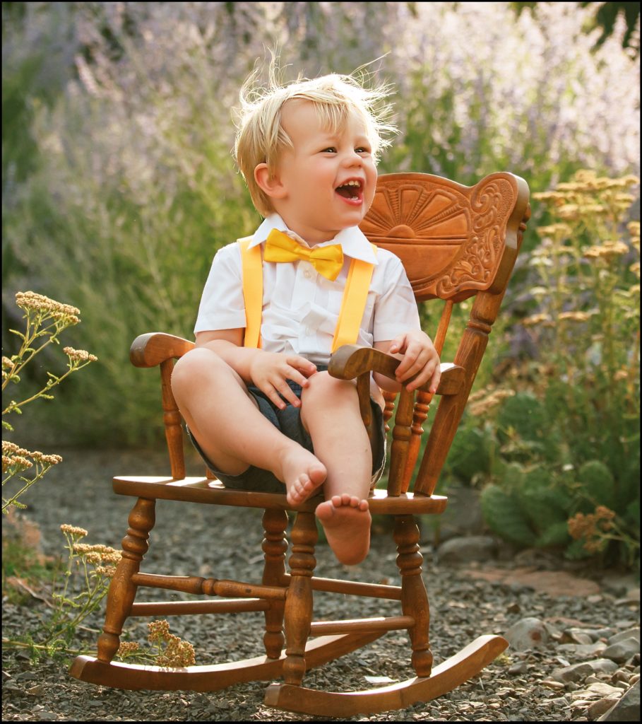 Little blonde boy in white shirt and yellow bow tie sits in a wooden rocking chair in a field in Washington. Ellensburg Family Photographer.