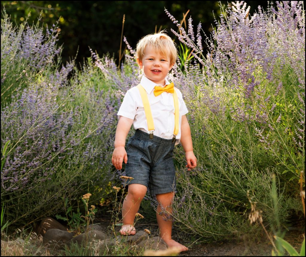 Little blonde boy in white shirt and yellow bow tie stands barefoot in a field of wild flowers. Ellensburg Family Photographer.