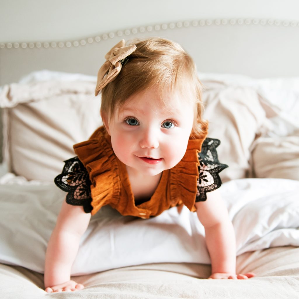 One year old baby girl in a rust colored dress on a bed taken by a family photographer in Ellensburg
