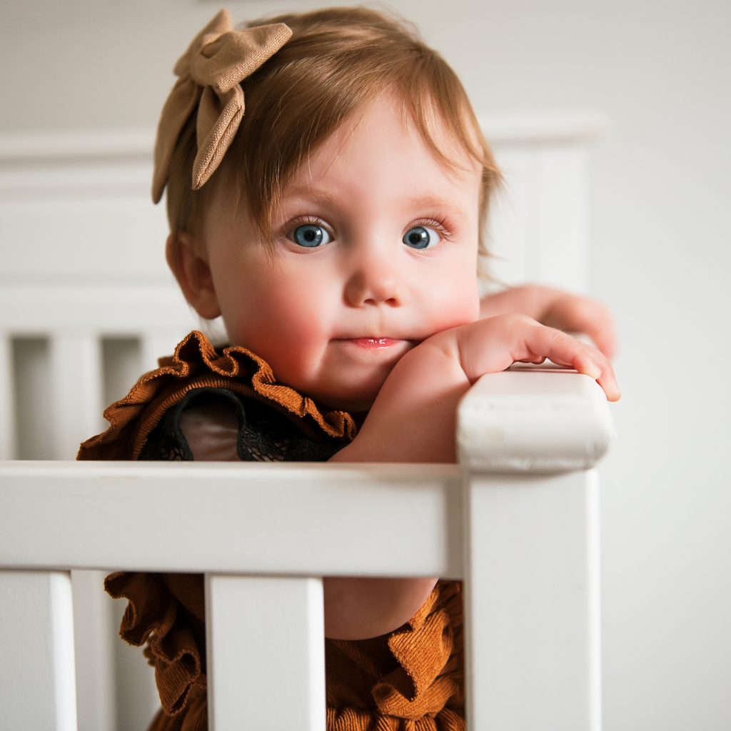 Family photographer in Ellensburg one year old stands in crib smiling at camera